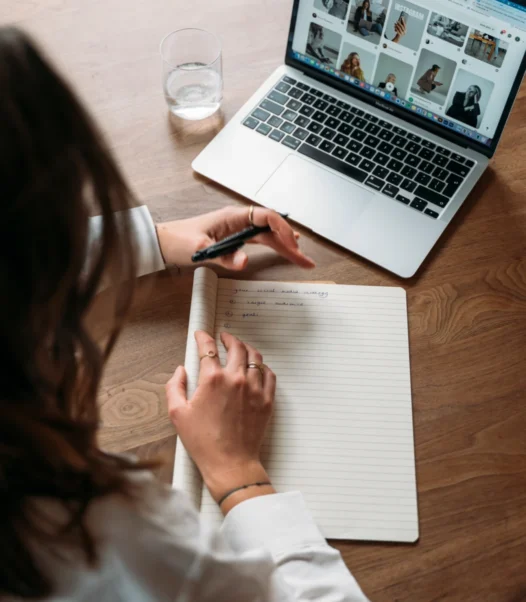 Het schrijven van een social media strategie op een notitieblok terwijl een laptop open staat met een Pinterest pagina die inspiratie biedt voor content voor Instagram, Facebook en LinkedIn.